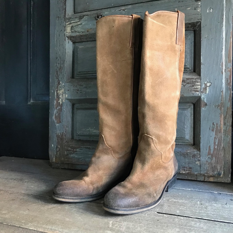 May include: A pair of knee-high, tan suede boots with a worn, distressed appearance. The boots have a slight heel and a dark brown leather accent at the top. The boots are standing on a wooden surface.