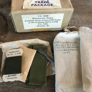 May include: A brown cardboard box with the text "TRADE PACKAGE" and "CC 2220 Housewives Packs, Tropical Areas, Green 1953 Pattn: A-78-Clothing-16171-CTSB P.I. 1966 Quantity 20". The box is surrounded by four white fabric bags, one of which contains a spool of green thread labeled "BARBOUR'S No. 35 - 2 CORD 50 YARDS ROTPROOFED".