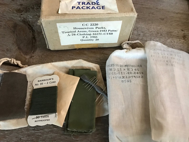 May include: A brown cardboard box with the text "TRADE PACKAGE" and "CC 2220 Housewives Packs, Tropical Areas, Green 1953 Pattn: A-78-Clothing-16171-CTSB P.I. 1966 Quantity 20". The box is surrounded by four white fabric bags, one of which contains a spool of green thread labeled "BARBOUR'S No. 35 - 2 CORD 50 YARDS ROTPROOFED".