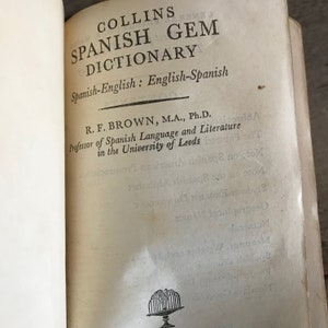 May include: A vintage book titled "Collins Spanish Gem Dictionary" with the subtitle "Spanish-English: English-Spanish". The book is authored by R. F. Brown, M.A., Ph.D., Professor of Spanish Language and Literature in the University of Leeds.