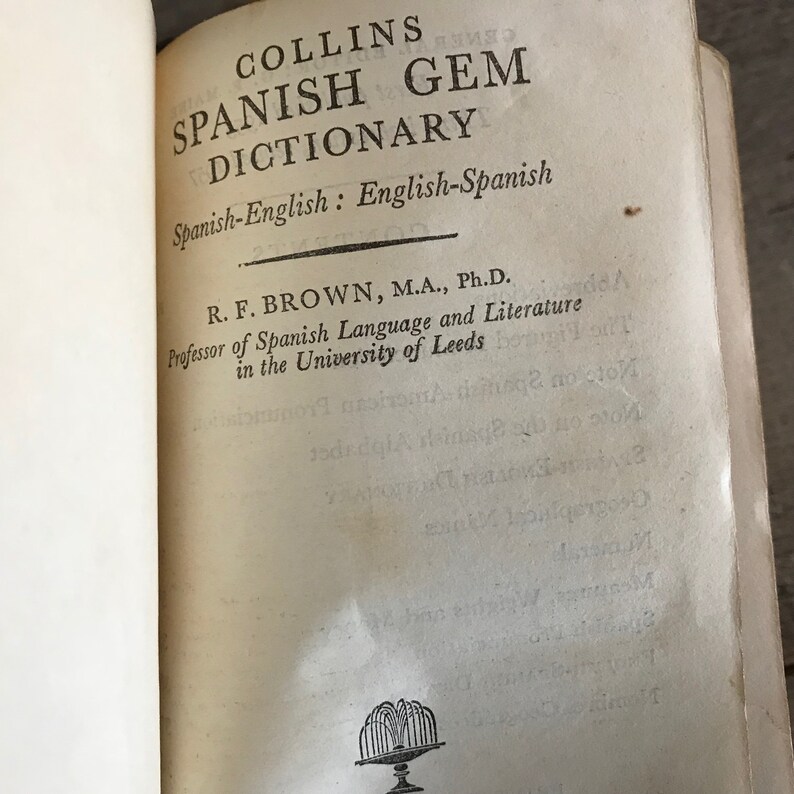 May include: A vintage book titled "Collins Spanish Gem Dictionary" with the subtitle "Spanish-English: English-Spanish". The book is authored by R. F. Brown, M.A., Ph.D., Professor of Spanish Language and Literature in the University of Leeds.