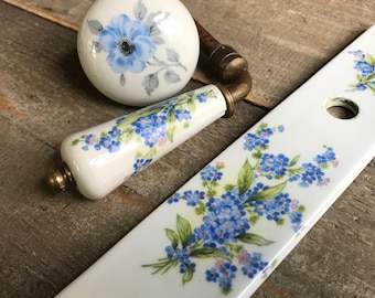 Könnte beinhalten: Ein Satz weißer Porzellanknopf für Türen mit floralem Design. Die Knöpfe zeigen blaue Vergissmeinnicht-Blumen und grüne Blätter. Die Knöpfe sind mit goldenen Beschlägen verziert.