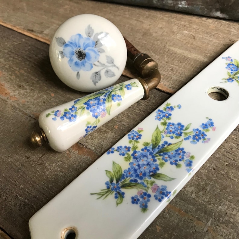 May include: A set of white porcelain door knobs with a floral design. The knobs feature blue forget-me-not flowers and green leaves. The knobs are accented with gold hardware.