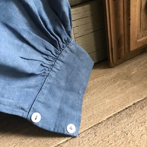 May include: Close-up of a blue linen shirt sleeve with a gathered cuff and two white buttons.