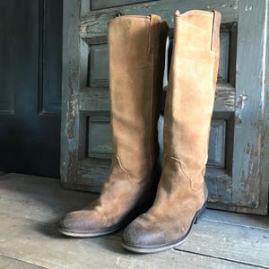 May include: A pair of knee-high brown suede boots. The boots have a worn, distressed appearance with a slight heel and a rounded toe. The boots are standing on a wooden surface in front of a weathered, blue-green door.