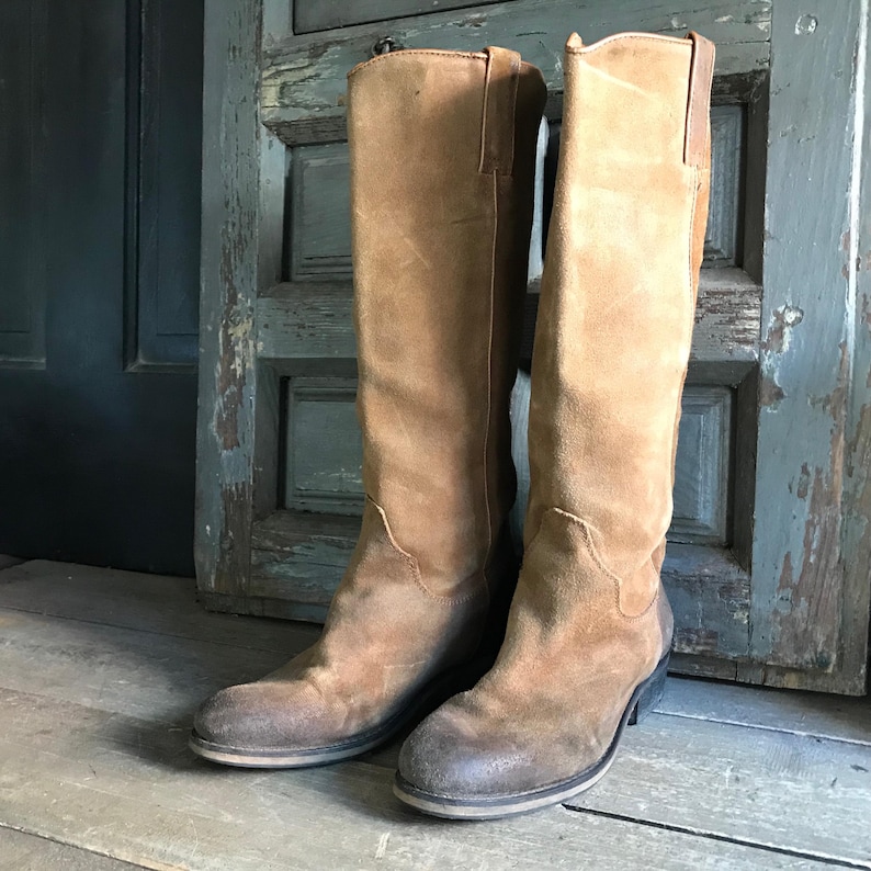 May include: A pair of knee-high brown suede boots. The boots have a worn, distressed appearance with a slight heel and a rounded toe. The boots are standing on a wooden surface in front of a weathered, blue-green door.
