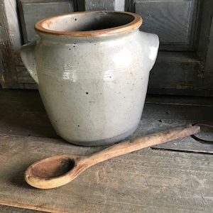May include: A light gray ceramic crock with a wide opening, a brown rim, and two side handles, accompanied by a long-handled wooden spoon with a dark, worn bowl and a leather loop attached to the end of the handle.