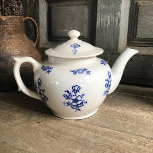 May include: White ceramic teapot with a lid, adorned with blue floral patterns. The teapot features a curved handle and spout. The lid has a small mushroom design. The teapot is sitting on a wooden surface.