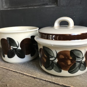 Puede incluir: Un juego de jarra de crema y azucarero de cerámica blanca con un patrón floral marrón y verde. La jarra tiene un pico y el azucarero tiene una tapa con una manija.