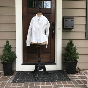 May include: A white, long-sleeved shirt with a ruffled collar and a button-up front. The shirt is displayed on a black stand in front of a wooden door.