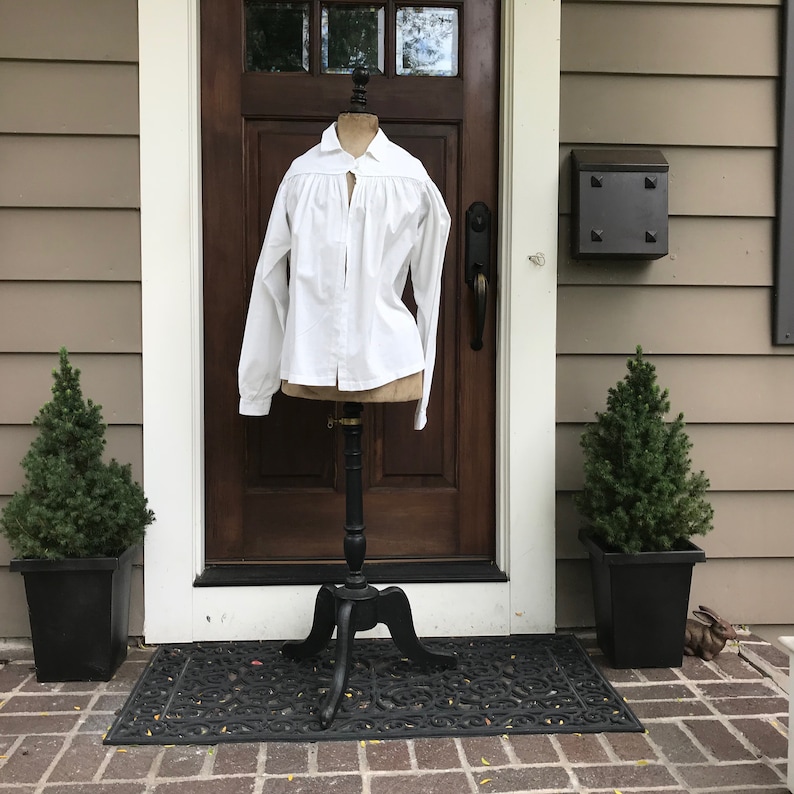 May include: A white, long-sleeved shirt with a ruffled collar and a button-up front. The shirt is displayed on a black stand in front of a wooden door.