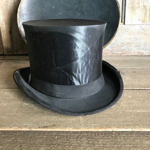 May include: A black top hat with a satin finish. The hat has a wide brim and a tall crown.
