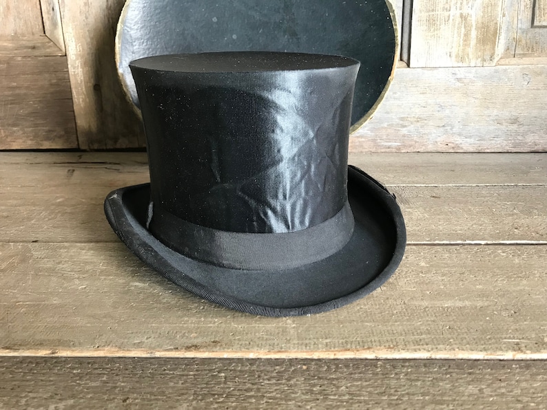 May include: A black top hat with a satin finish. The hat has a wide brim and a tall crown.