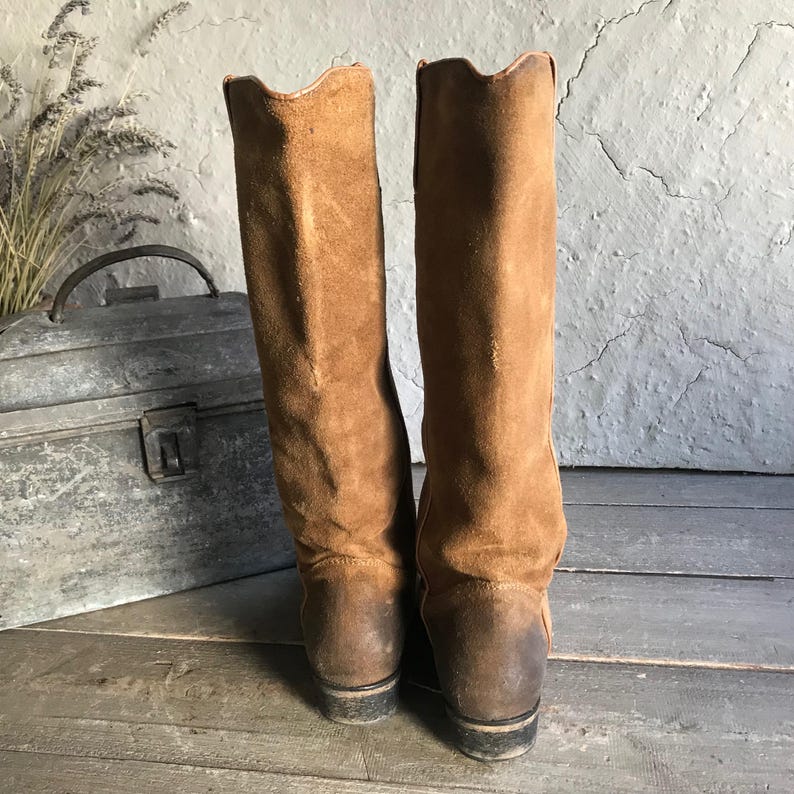 May include: A pair of tall, brown suede boots with a distressed appearance. The boots have a classic design with a slightly curved top edge and a stacked heel. The boots are set against a rustic background.