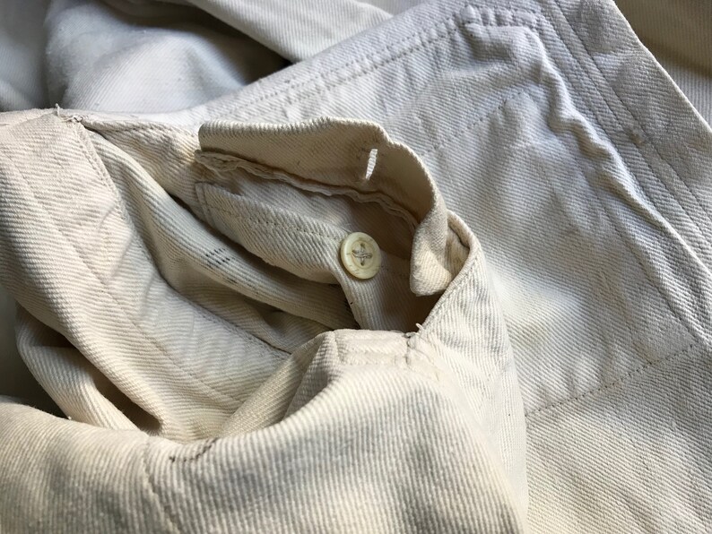 K&ouml;nnte beinhalten: Nahaufnahme eines cremefarbenen Jeansjackenkragens mit Knopfverschluss. Der Stoff hat ein strukturiertes Aussehen mit sichtbaren Nahtdetails. Der Knopf ist hellbeige.