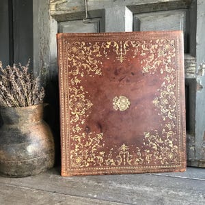 Może przedstawiać: Duża, antyczna, brązowa, skórzana oprawa książki z bogatymi złotymi wzorami kwiatowymi. Okładka posiada centralny herb i dekoracyjne obramowania. Książka umieszczona jest na zwietrzałej drewnianej powierzchni, obok rustykalnego wazonu z suszoną lawendą.