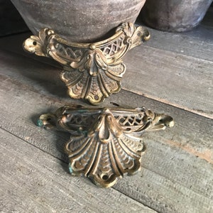 May include: Two antique brass furniture corner brackets. The brackets have an ornate, symmetrical design with a central floral motif and openwork detailing. The brass has a warm, aged patina with hints of green oxidation, suggesting age and use. The brackets are displayed on a weathered wooden surface.
