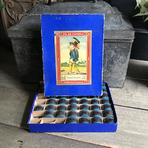 Könnte beinhalten: Eine blaue Box mit einer Cartoon-Illustration eines Mannes, der einen Spazierstock hält. Die Box enthält 24 kleine blaue Kapseln mit Garn. Auf der Box steht "Fil en Capsule" und "50 Metres Garantis".