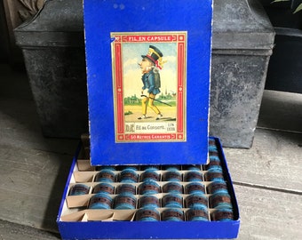 May include: A blue box with a cartoon illustration of a man holding a walking stick. The box contains 24 small blue capsules of thread. The box reads "Fil en Capsule" and "50 Metres Garantis".