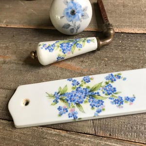 May include: A set of three white ceramic door knobs with a floral design. The knobs feature a blue and green floral pattern with forget-me-not flowers. The knobs are attached to a brass base.