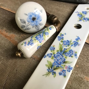 May include: A set of white porcelain door knobs with a floral design. The knobs feature blue forget-me-not flowers with green leaves.