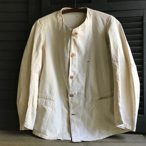 K&ouml;nnte beinhalten: Cremefarbene Arbeitsjacke mit Stehkragen und Holzkn&ouml;pfen. Die Jacke hat zwei Vordertaschen und h&auml;ngt an einem Holzkleiderb&uuml;gel. Der Hintergrund umfasst einen dunklen Holzladen und einen Keramikkrug.