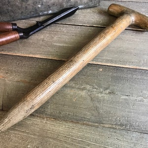May include: A long, wooden handle for a garden tool. The handle is light brown with a weathered finish and has a T-shaped top.
