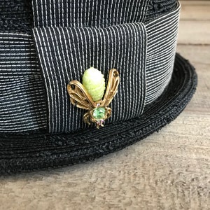 May include: A black and white striped hat with a gold bee pin with a green gem. The bee pin has a light green body and gold wings.