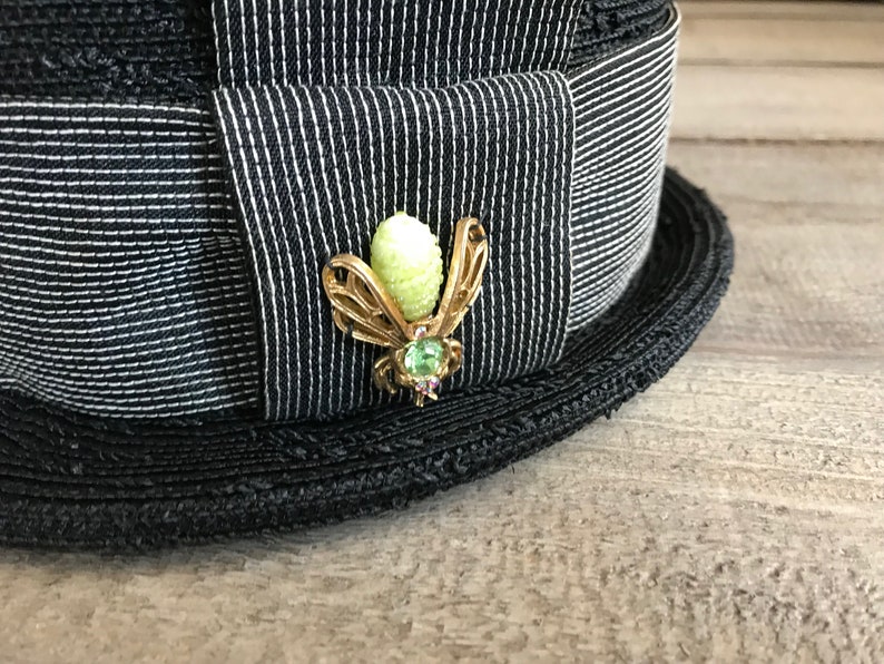 May include: A black and white striped hat with a gold bee pin with a green gem. The bee pin has a light green body and gold wings.