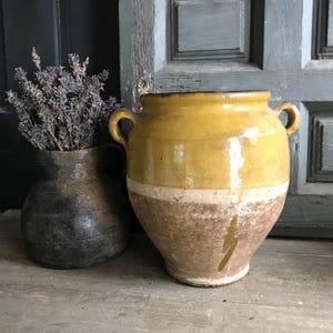 May include: Two antique ceramic vases. One is a dark, textured vase holding dried lavender. The other is a large, yellow and brown vase with two handles and a white band around the middle. The vases are on a wooden surface.
