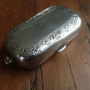 Puede incluir: Una caja rectangular de plata con bisagras y un diseño ornamentado. La caja tiene un pequeño lazo en un extremo.