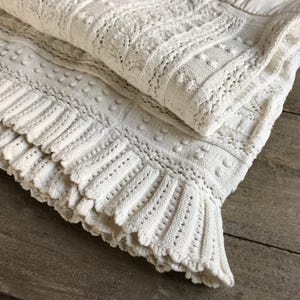 May include: Cream-colored knitted blanket with a textured design. The blanket features a variety of patterns, including ribbed sections, dotted details, and a scalloped edge. The blanket is folded, showcasing its intricate craftsmanship and soft texture.