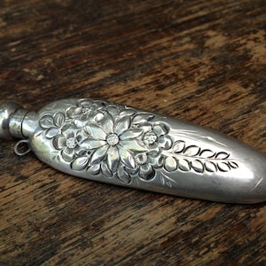 Puede incluir: Una botella alargada de plata, con relieve floral, con tapa con bisagra. La botella está decorada con un diseño floral en relieve y tiene un acabado liso y pulido.