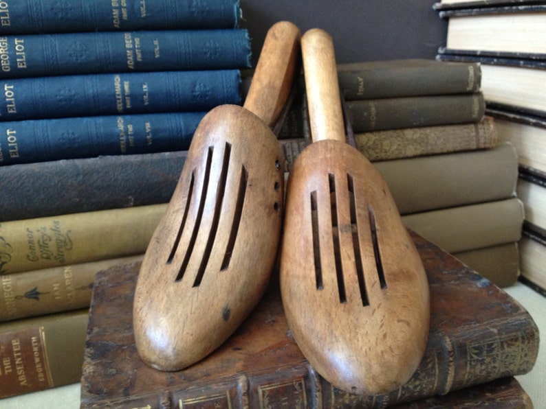 May include: Two wooden shoe trees with slotted fronts, resting on a stack of antique books. The shoe trees are light brown with a smooth finish. The books have aged covers in shades of blue, green, and brown.