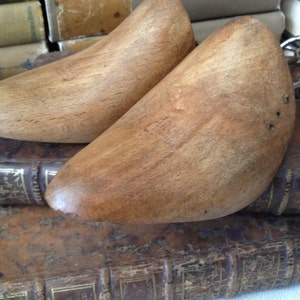 May include: A pair of wooden shoe trees with a metal connecting piece. The shoe trees are brown and have a smooth, polished finish. They are resting on top of a stack of old books.