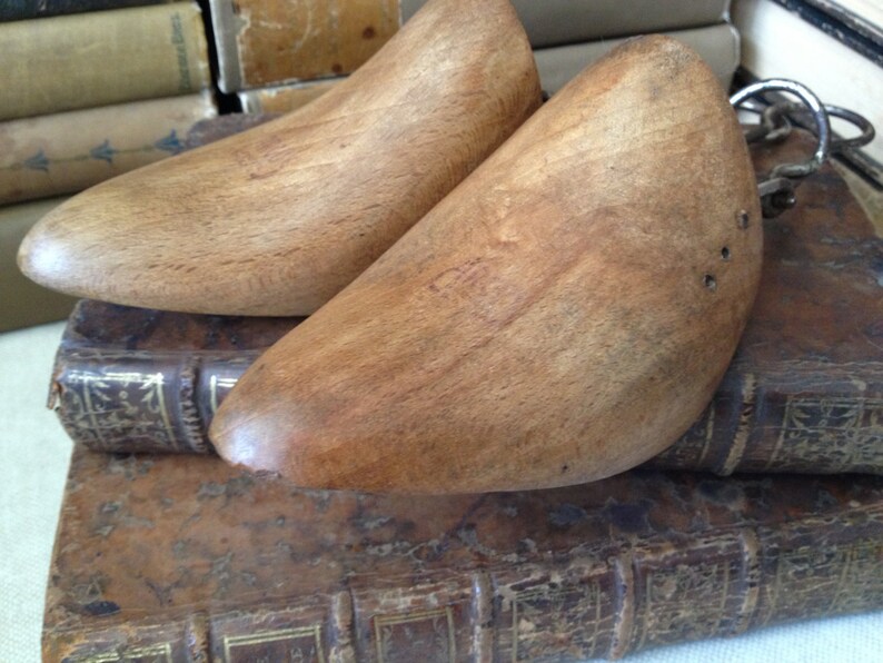 May include: A pair of wooden shoe trees with a metal connecting piece. The shoe trees are brown and have a smooth, polished finish. They are resting on top of a stack of old books.
