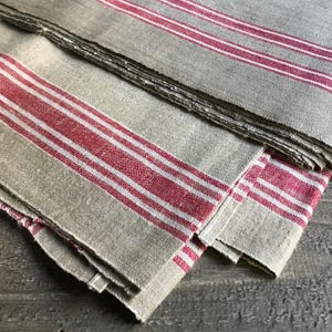 Pode incluir: Uma pilha de guardanapos de linho bege com listras vermelhas e brancas arrojadas. Os guardanapos têm uma aparência rústica e texturizada, sugerindo um estilo vintage ou de fazenda. As listras vermelhas são espaçadas uniformemente no tecido.