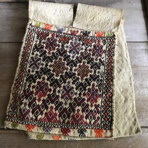 May include: A white fabric pouch with a geometric pattern embroidered in red, orange, purple, and black thread. The pouch is folded in half and has a raw edge.