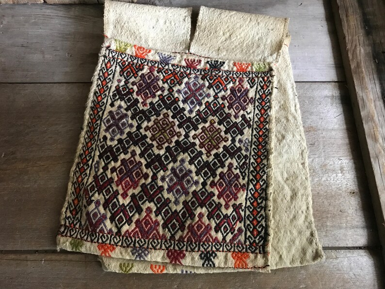 May include: A white fabric pouch with a geometric pattern embroidered in red, orange, purple, and black thread. The pouch is folded in half and has a raw edge.