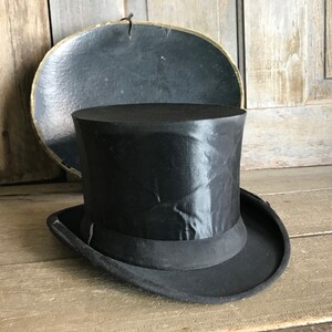 May include: A black top hat with a satin finish and a black ribbon band. The hat is sitting on a wooden surface.
