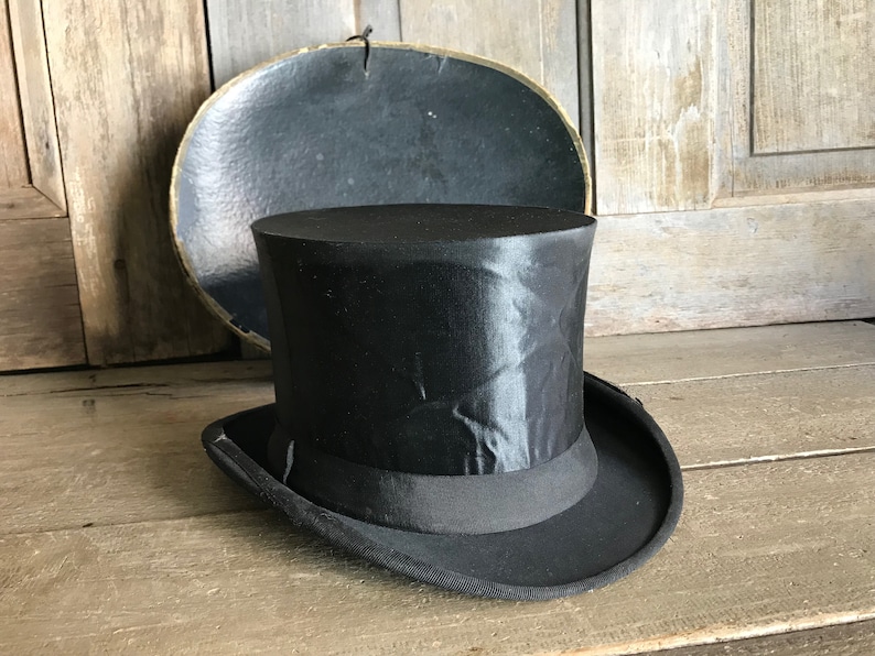 May include: A black top hat with a satin finish and a black ribbon band. The hat is sitting on a wooden surface.