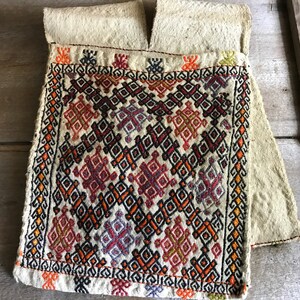 May include: A beige fabric pouch with a geometric pattern embroidered in red, orange, black, and purple thread. The pouch has a flap at the top and is folded over.