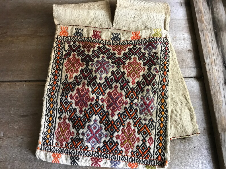 May include: A beige fabric pouch with a geometric pattern embroidered in red, orange, black, and purple thread. The pouch has a flap at the top and is folded over.