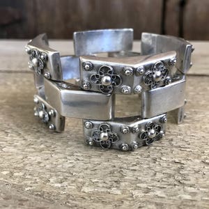 May include: A silver bracelet with a geometric design. The bracelet is made of interlocking pieces with floral accents.