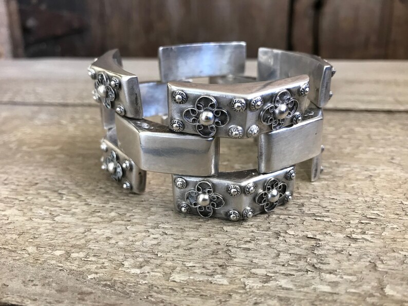 May include: A silver bracelet with a geometric design. The bracelet is made of interlocking pieces with floral accents.