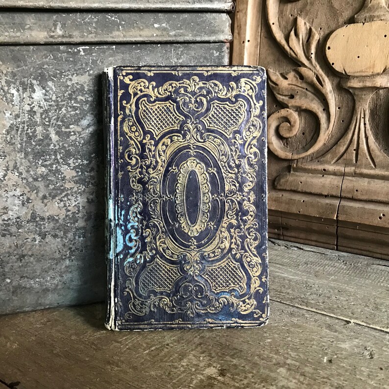 May include: A vintage book with a dark blue cover and gold ornate design. The cover features a central oval shape with a gold border and intricate floral patterns.