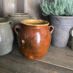 May include: A large, brown earthenware crock with a yellow glaze on the rim. The crock has a rounded body and two handles. It is sitting on a wooden surface.