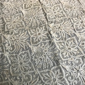 以下が含まれることがあります： 繊細で複雑な刺繍の花と葉の模様が施された白い綿の生地。デザインはグリッドパターンで繰り返され、対称的でエレガントな外観を作り出しています。