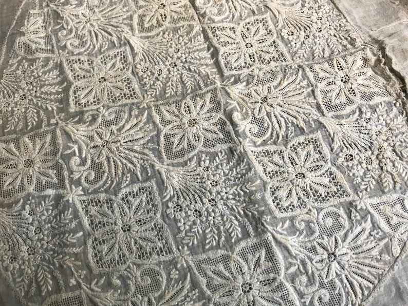 以下が含まれることがあります： 繊細で複雑な刺繍の花と葉の模様が施された白い綿の生地。デザインはグリッドパターンで繰り返され、対称的でエレガントな外観を作り出しています。