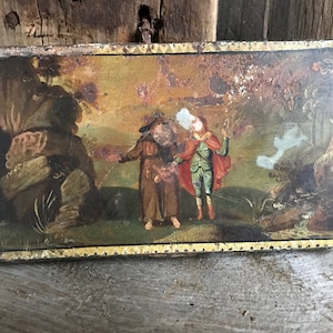 May include: A vintage tin box with a painted scene of three figures in a landscape. The figures are wearing colorful clothing and are standing in a field with trees and a stream. The box is decorated with a gold border.
